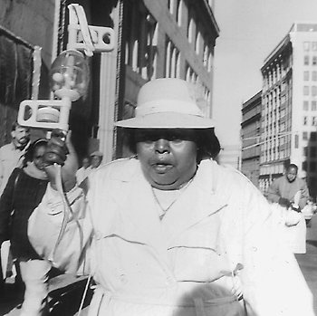 Véronique Jean-Pierre nan yon manifestasyon demokratik nan Tremont Street, Downtown Boston, an 1990. Gen moun ki rele manmzèl «Mandan Lavalas» paske li toujou defann avèk anpil pasyon revandikasyon mouvman rezistans demokratik la kont koudeta anti-popilè Cedras la, avèk yon adorasyon san kondisyon pou pè Titid. —fotografye pa David Henry.